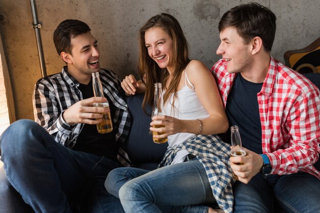 Giovani felici seduti sul divano, bere birra, chiudere le mani tostare, divertirsi, festa a casa di amici, compagnia hipster insieme, due uomini una donna, sorridente, positivo, rilassato, uscire, ridere