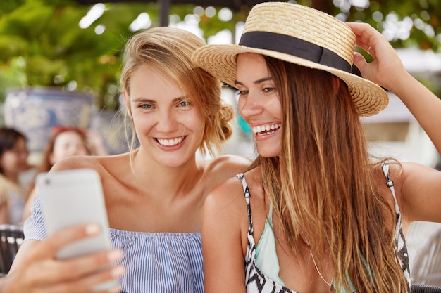 Giovani femmine felici guardano video interessanti su smartphone o fanno selfie, hanno uno sguardo deliziato, riposano insieme alla caffetteria all'aperto nella località turistica. Persone, relazioni e concetto di riposo estivo