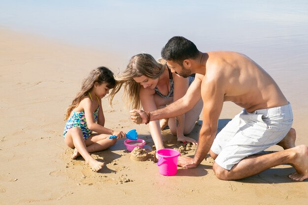 Giovani genitori e dolce bambina che giocano insieme sulla spiaggia, costruendo costruzioni di sabbia con pala giocattolo, secchio e ciotola