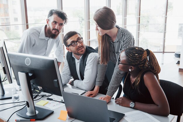 Giovani impiegati che si siedono nell'ufficio alla tavola e che per mezzo di un computer portatile, un concetto di riunione di "brainstorming" del lavoro di gruppo.