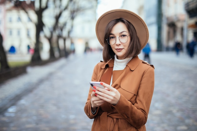 Giovani in cappotto marrone che manda un sms sulla via di autunno dello smartphone