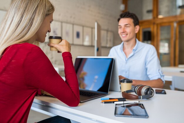 Giovani moderni seduti a tavola faccia a faccia, lavorando al computer portatile in ufficio di co-working