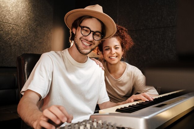 Giovani musicisti allegri che guardano felicemente nella fotocamera che registrano la canzone sulla tastiera del pianoforte per il nuovo album musicale in uno studio moderno