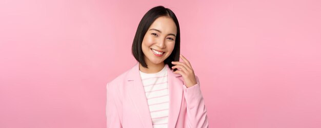 Giovani professionisti La commessa asiatica sorridente della donna di affari in vestito che guarda fiduciosa alla macchina fotografica che posa contro lo sfondo rosa