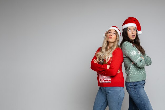 Giovani ragazze di natale abbastanza sorprese in cappelli di babbo natale e maglioni invernali in posa su sfondo grigio studio con spazio copia