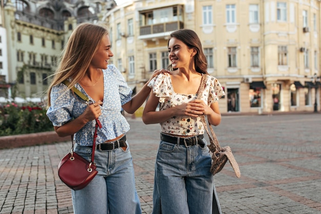 Giovani ragazze felici in pantaloni di jeans e camicette floreali alla moda camminano fuori a parlare e sorridono Ritratto di fidanzate bionde e brune posano nella piazza della città