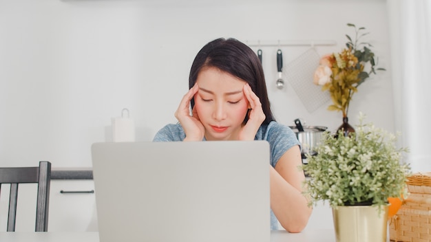 Giovani record asiatici della donna di affari di entrate e spese a casa. Signora preoccupata, seria, stress durante l'utilizzo del budget record del laptop, fiscale, documento finanziario che lavora nella cucina moderna a casa.