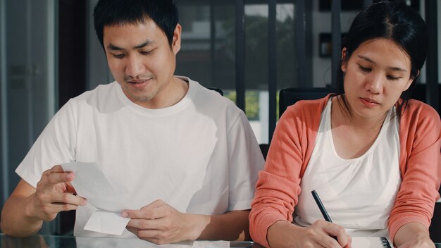 Giovani record incinti asiatici delle coppie delle entrate e delle spese a casa. Mamma e papà felici utilizzando il budget record del computer portatile, tasse, documento finanziario, commercio elettronico che lavora nel salotto di casa.