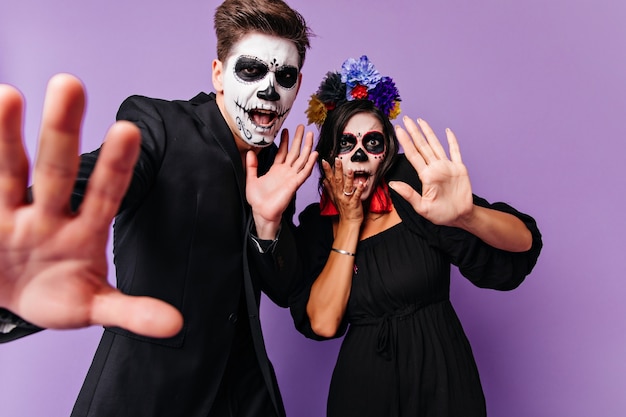 Giovani spaventati in abiti di halloween che stanno insieme su sfondo viola. Foto dell'interno di una coppia europea entusiasta che scherza in costumi muertos.