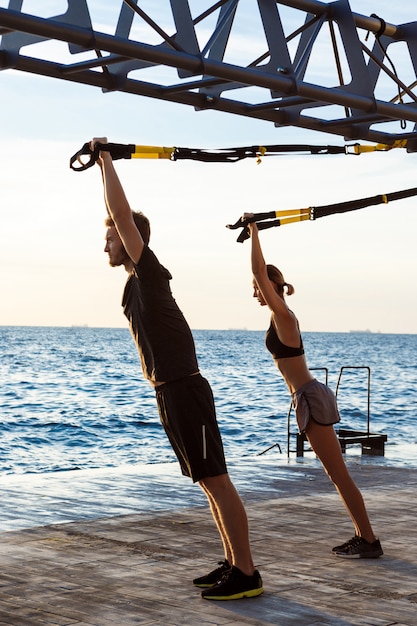 Giovani sportivi che si allenano con trx vicino al mare al mattino.