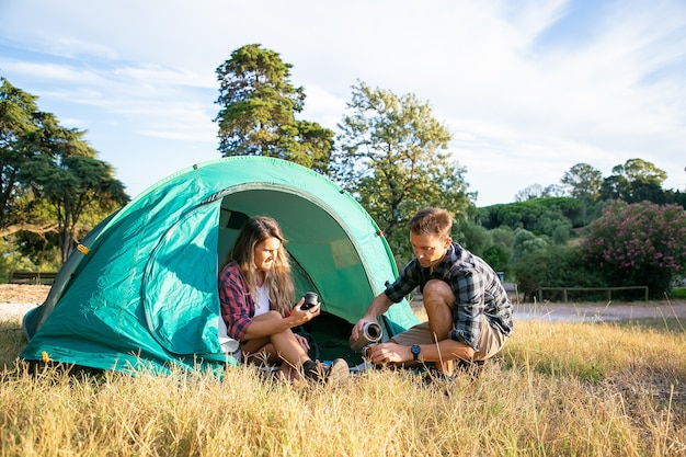 Giovani turisti caucasici in campeggio sul prato e seduti in tenda. Coppia felice di bere il tè dal thermos e rilassarsi sulla natura insieme. Concetto di turismo, avventura e vacanze estive con lo zaino in spalla