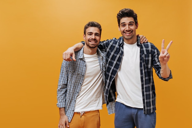 Giovani uomini allegri in camicie blu a quadri, magliette bianche e pantaloni colorati posano sulla parete arancione di ottimo umore e sorridono.