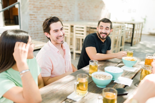 Giovani uomini e donne felici che si divertono seduti insieme in un bar all'aperto. Amici allegri che parlano e che bevono birra