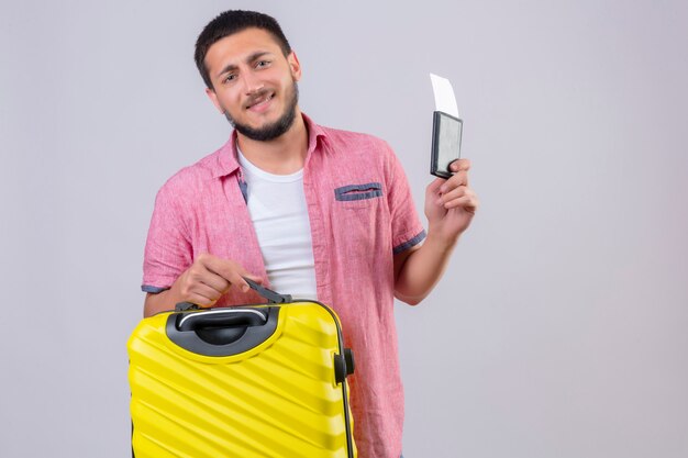 Giovani valigie e biglietti aerei bei della tenuta del tipo del viaggiatore che esaminano macchina fotografica con il sorriso sicuro sul fondo bianco controllante felice e positivo del fronte