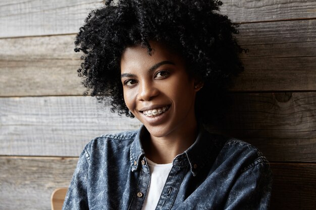 Gioventù e felicità. Bellezza e moda. Chiuda sul ritratto di giovane donna africana attraente felice con le parentesi graffe che gode del suo tempo libero all'interno del caffè moderno, vestito in camicia di jeans alla moda