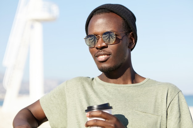 Gioventù, stile di vita moderno e concetto di felicità. Giovane viaggiatore afroamericano sembrante d'avanguardia attraente in occhiali da sole con lenti a specchio che si rilassano sul banco nella stazione turistica, mangiando caffè e respirando aria di mare