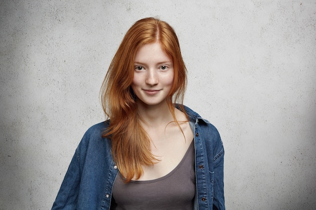 Giovinezza e felicità. Vista ravvicinata della bella donna adolescente caucasica con lunghi capelli rossi e lentiggini, vestita casualmente, in piedi contro il muro grigio vuoto.