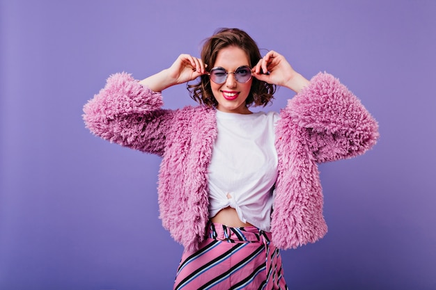 Glamour giovane donna con i capelli ondulati scuri che tocca i suoi occhiali alla moda. Ritratto dell'interno di piacevole ragazza caucasica in soffice giacca di pelliccia isolato sulla parete viola.