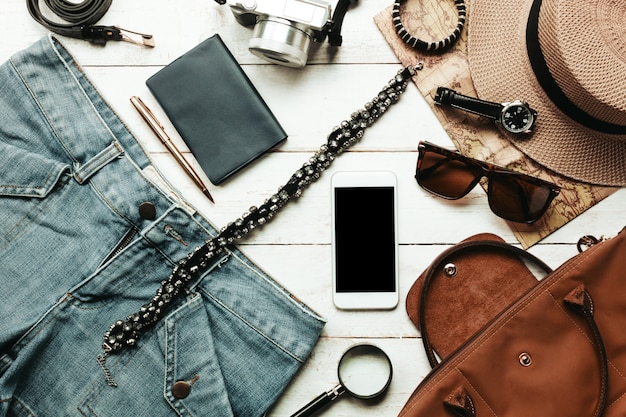 Gli accessi di vista superiore per viaggiare con il concetto di abbigliamento femminile. Cellulare bianco, orologio, borsa, cappello, mappa, fotocamera, collana, pantaloni e occhiali da sole su tavola di legno bianco.