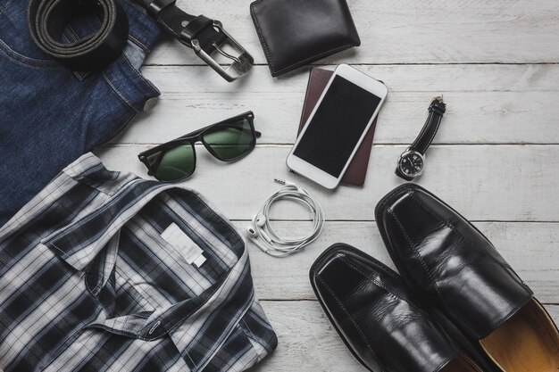 Gli accessi di vista superiore per viaggiare con il concetto di abbigliamento uomo. shirt, jean, telefono cellulare su background.watch di legno, occhiali da sole e scarpe sul tavolo di legno.