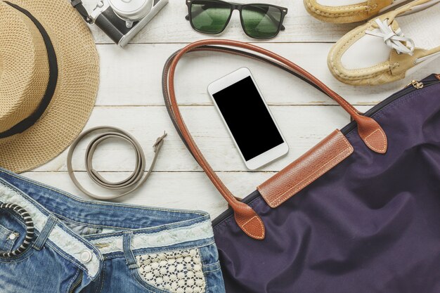 Gli accessori accessibili in alto per viaggiare con le donne vestono il cellulare, la cintura, la borsa, il cappello, la macchina fotografica, la collana, i pantaloni e gli occhiali da sole sul tavolo di legno bianco.