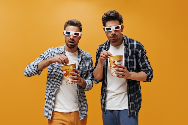 Gli amici guardano film con occhiali 3D su sfondo arancione Gli uomini brunet in camicie a scacchi mangiano popcorn e posano isolati