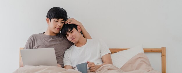 Gli omosessuali asiatici si accoppiano facendo uso del computer portatile del computer e bevendo il caffè a casa moderna. Il giovane maschio dell'amante dell'Asia felice si rilassa insieme dopo il risveglio, guardando il film che si trova sul letto in camera da letto a casa nella mattina