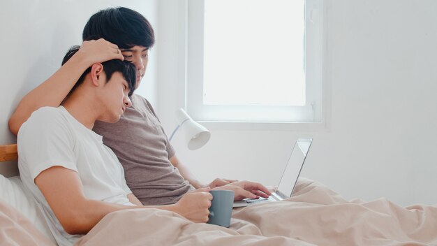 Gli omosessuali asiatici si accoppiano facendo uso del computer portatile del computer e bevendo il caffè a casa moderna. Il giovane maschio dell'amante dell'Asia felice si rilassa insieme dopo il risveglio, guardando il film che si trova sul letto in camera da letto a casa nella mattina