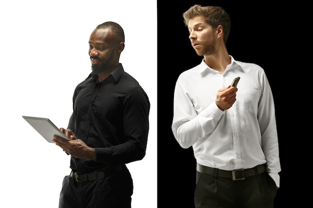 Gli uomini afro e caucasici felici di successo. Coppia mista con gadget. Immagine dinamica di modelli maschili in studio bianco e nero. Concetto di emozioni facciali umane.