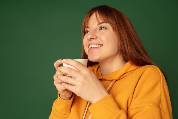 Godendo di caffè tè ritratto di donna caucasica su sfondo verde studio con copyspace Bellissimo modello femminile in felpa gialla Concetto di emozioni umane espressione facciale ad moda