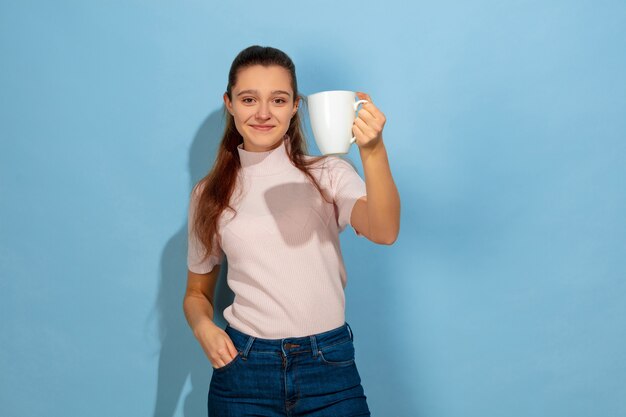 Godendo di caffè, tè, sembra calmo. Ritratto della ragazza teenager caucasica su priorità bassa blu. Bellissima modella in abbigliamento casual. Concetto di emozioni umane, espressione facciale, vendite, annuncio. Copyspace. Sembra felice.