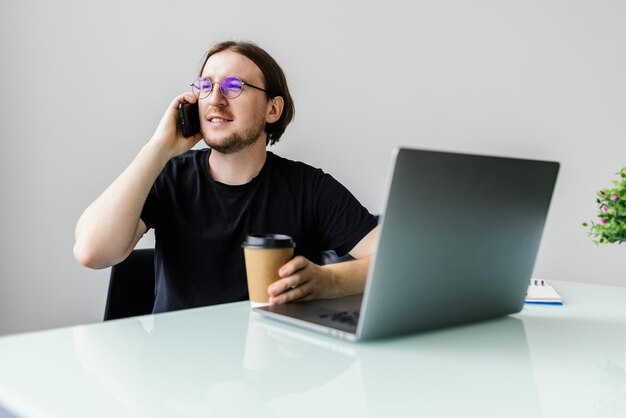 Godersi una buona giornata di lavoro Giovane fiducioso che lavora su un laptop e parla al telefono cellulare mentre è seduto al suo posto di lavoro in ufficio