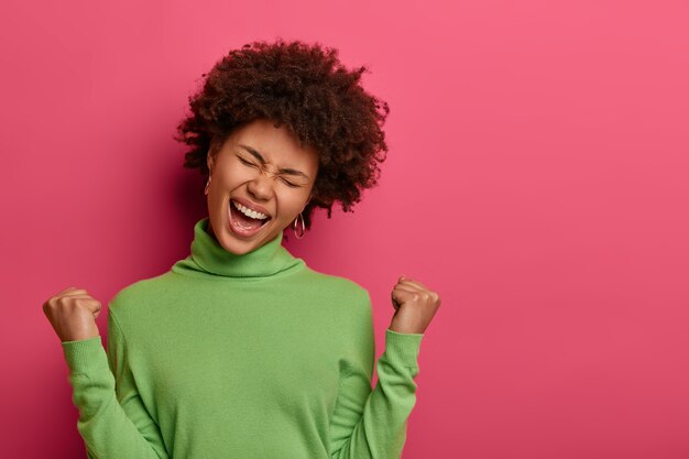 Grande successo e concetto di successo. Felice donna afroamericana stringe i pugni, fa un gesto di vittoria, urla sì, inclina la testa, indossa un dolcevita verde