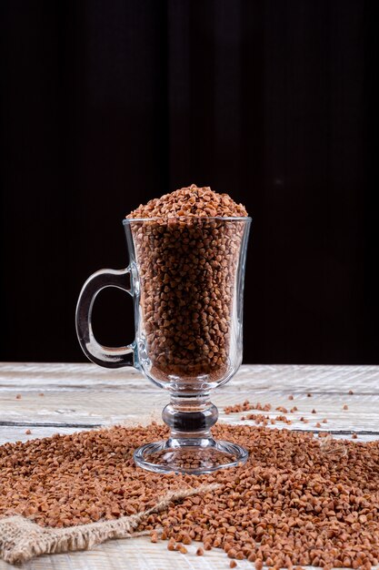 Grano saraceno di vista laterale in tazza con grano saraceno sotto esso su fondo di legno e nero bianco. spazio orizzontale per il testo