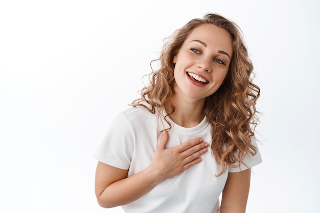 Grata ragazza bionda sorridente, tenendo la mano sul cuore e guardando da parte il testo promozionale con volto grato e compiaciuto, in piedi sul muro bianco