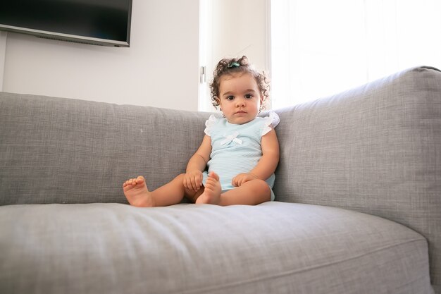 Grave bambina dai capelli ricci scuri che indossa un panno blu pallido, seduta sul divano grigio a casa, a. Kid a casa e il concetto di infanzia