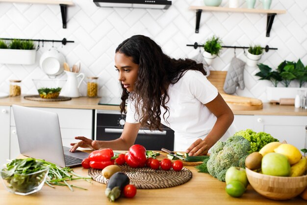 Grave bella mulatta sta guardando sullo schermo del laptop sulla cucina moderna sul tavolo pieno di frutta e verdura, vestita con una maglietta bianca