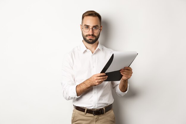 Grave datore di lavoro che guarda l'obbiettivo durante la lettura di documenti negli appunti, avendo colloquio di lavoro, in piedi