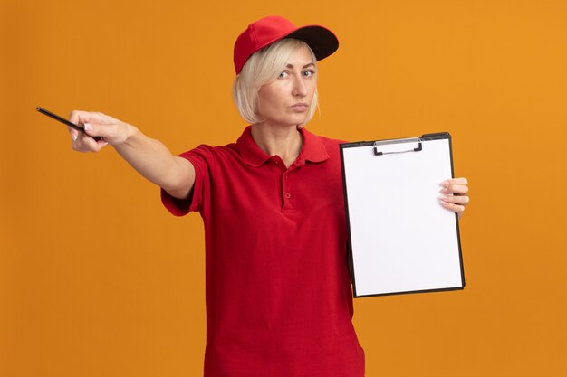 Grave donna di mezza età bionda consegna in uniforme rossa e berretto che tiene appunti guardando la parte anteriore che punta a lato con la matita isolata sulla parete arancione
