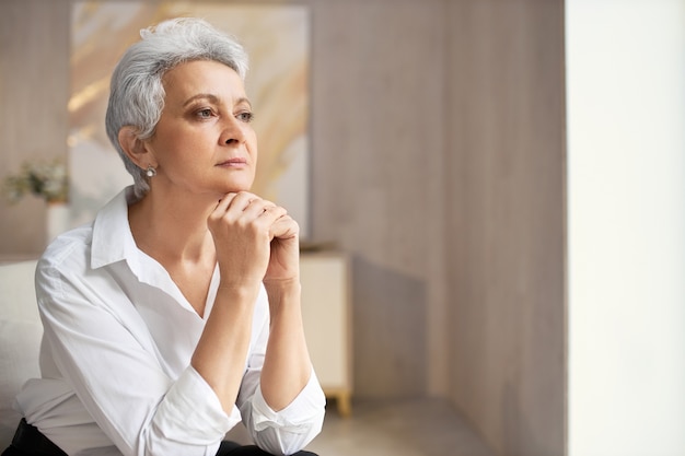 Grave elegante donna in pensione con acconciatura corta in posa in casa con le mani sotto il mento, guardando lontano con espressione facciale pensierosa, pensando a qualche idea o decisione