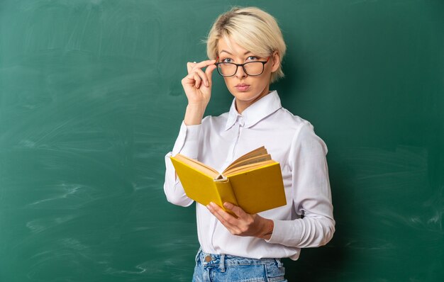 Grave giovane insegnante di sesso femminile bionda con gli occhiali in aula in piedi nella vista di profilo davanti alla lavagna che tiene il libro afferrando gli occhiali guardando la fotocamera con lo spazio della copia