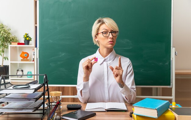 Grave giovane insegnante di sesso femminile bionda con gli occhiali seduto alla scrivania con strumenti di scuola in aula guardando la telecamera che mostra un piccolo quadrato a forma di numero cinque alzando il dito