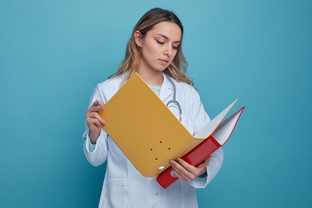 Grave giovane medico femminile che indossa abito medico e stetoscopio intorno al collo tenendo la penna e le cartelle guardando uno aperto