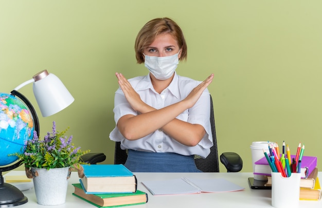 Grave giovane studentessa bionda che indossa una maschera protettiva seduta alla scrivania con gli strumenti della scuola che guarda l'obbiettivo che non fa alcun gesto isolato sul muro verde oliva