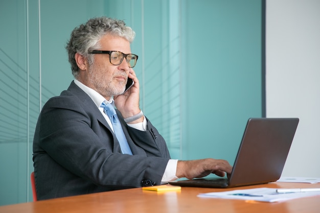 Grave imprenditore fiducioso in tuta e occhiali parlando al telefono cellulare, lavorando al computer in ufficio, utilizzando il computer portatile al tavolo con diagrammi di carta