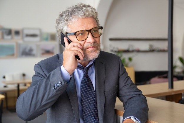 Grave imprenditore maturo impegnato con gli occhiali, parlando al telefono cellulare, in piedi al co-working, appoggiato sulla scrivania, guardando lontano