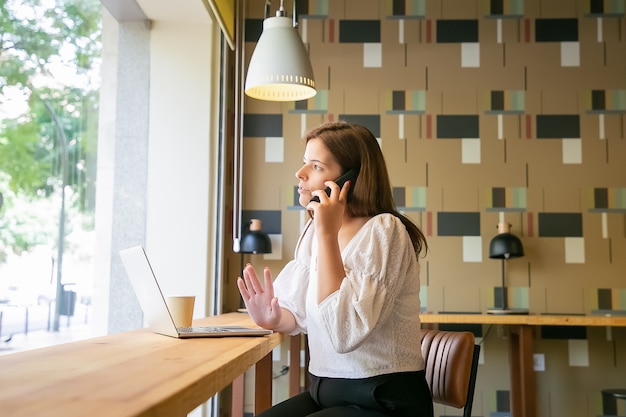 Grave professionista freelance parlando al cellulare mentre è seduto alla scrivania con laptop e caffè nello spazio di co-working