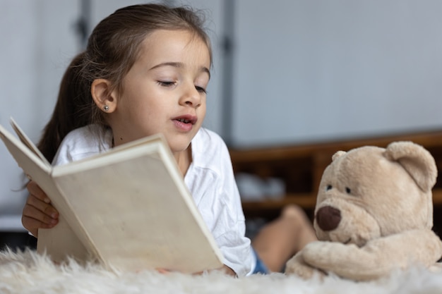 Graziosa bambina a casa, sdraiata sul pavimento con il suo giocattolo preferito e legge un libro.