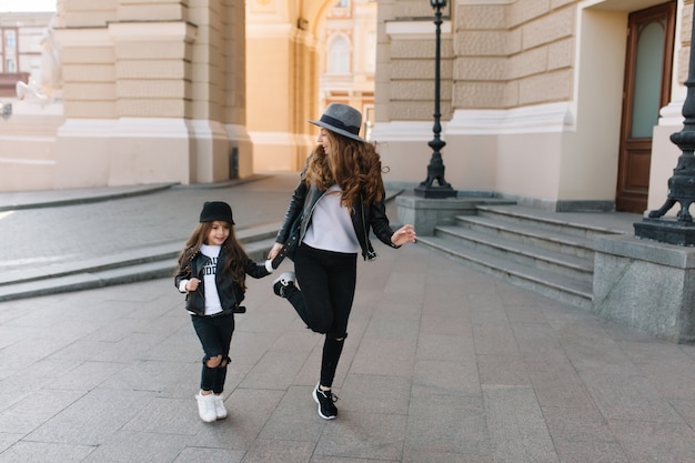 Graziosa giovane donna riccia in jeans skinny neri divertente ballare per strada vicino alla bambina gioiosa.