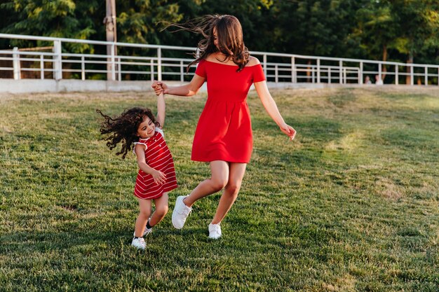 Graziosa giovane signora in breve vestito rosso che tengono le mani con la figlia. Foto a figura intera all'aperto della madre e del bambino che giocano nel parco.